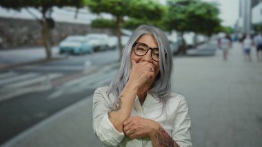 Arka planda arabalar ve ağaçlarla dolu bir caddede gülümseyen kır saçlı son sınıf öğrencisi kadın neşeli ve sıradan bir şehir manzarası sergiliyor..