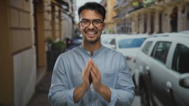Mavi gömlekli genç adam gündüz vakti şehir binaları boyunca park etmiş arabalarla dolu caddede heyecanını dile getiriyor..