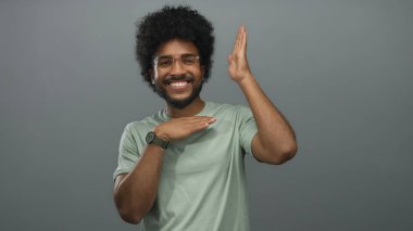 Gözlüklü genç adam gri bir duvara karşı neşeli jestler yapıyor, pozitif ve enerjik ifade ediyor, günlük kıyafetler giyiyor ve parlak bir şekilde gülümsüyor..