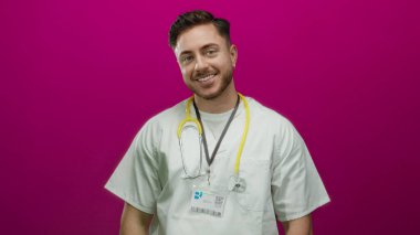 Steteskop ve sakallı, gülümseyen genç erkek doktor canlı pembe bir arka plana karşı duruyor, tıbbi bir ortamda profesyonel bakım ve neşeli bir tavır yakalıyor..