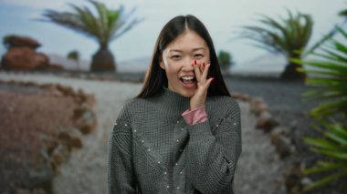 Parkta ağaçlar ve taşlarla gülümseyen, süveter giyen, huzurlu bir ortamda neşe ve doğa taşıyan bir kadın..