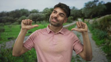 Yeşil tarlalar tarafından çerçevelenmiş bir orman yolu boyunca pembe bir polo tişörtle adam gülümser ve her iki başparmak kendine işaret eder; gurur.