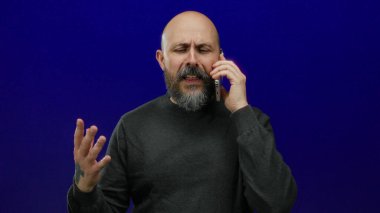Kel ve sakallı bir adam cep telefonuyla izole edilmiş mavi bir arkaplan karşısında konuşuyor koyu renk bir gömlek giyiyor ve el hareketleriyle kaygılarını açıkça ifade ediyor.