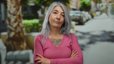 Gri saçlı yaşlı kadın açık havada kollarını kavuşturarak poz veriyor. Canlı bir şehir sahnesinde dövmesini gösteriyor..