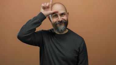 Kel kafalı, sakallı, siyah tişörtlü, komik yüzlü, parmağıyla işaret eden, kahverengi duvarlı ezik işareti yapan bir adam.