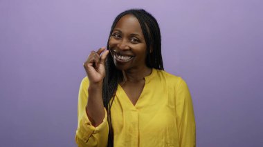 Sarı gömlekli bir kadın mor arka planda küçük bir jest yapıyor, özgüvenle gülümsüyor, pozitif duygu ve ifadeyi tasvir ediyor..