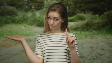 Genç beyaz kadın, yeşil bir parkta şüpheli görünürken avuç içi açık bir şekilde işaret ediyor..