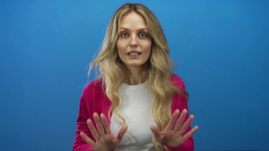 Elleri mavi bir arkaplanda, pembe bir kazak ve beyaz bir gömlek giymiş sakin bir şekilde duran ve izole bir sahneye odaklanan bir kadın..