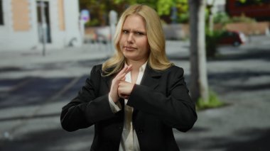 Şehirdeki genç sarışın kadın, şehir mimarisi ve arka planda bulanık manzarayla çevrili ciddi bir ifadeyle kavga jesti yapıyor..