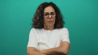 Orta yaşlı İspanyol kadın yeşil stüdyoda gözlük takarken kollarını kavuşturuyor ve ciddi şekilde kaşlarını çatıyor; reddediyor..