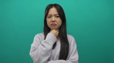 İzole edilmiş yeşil arka plan yüzünden boğazı ağrıyan bir kadın, derin derin düşünüyor, ensesine dokunuyor, sıradan bir kazak giyiyor..