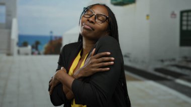 Kadın kentin sokaklarında kendini kucaklıyor, modern gözlüklerden ve güneşli gökyüzünün altında gündelik kıyafetlerden tatmin ve sakinliğini ifade ediyor..