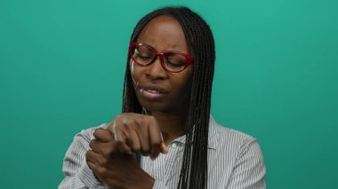 Gözlüklü bir kadın, yeşil bir duvara karşı bileklerini inceliyor. Odaklanmış bir ortamda tefekküre ya da endişeye işaret ediyor..