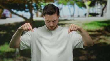 Beyaz tişörtlü beyaz sakallı genç beyaz bir adam parkın arka planında ağaçlar ve yeşillikler olan bir yeri işaret ediyor..