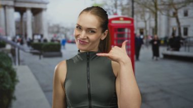Kadın, şehirdeki kırmızı telefon kulübesinden önce baş parmak ve parmak ucuyla telefon jesti yapıyor..