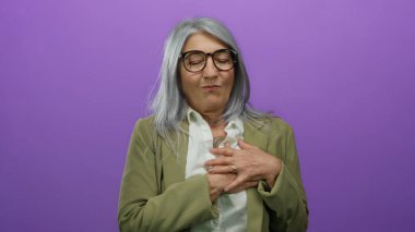 Gri saçlı ve gözlüklü yaşlı bir kadın, izole edilmiş mor arka plana karşı göğsünü sıkıyor, sağlıkla ilgili endişelerini ve duygularını iletiyor..