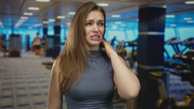 Kafkasyalı genç bir kadın spor salonunun içinde acı dolu bir ifadeyle boynunu tutuyor, arkasında koşu bantları var; rahatsız edici..