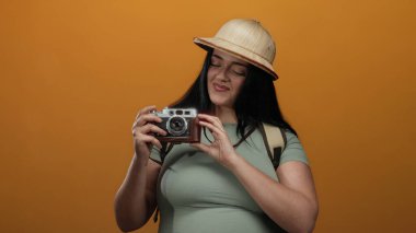 Kameralı kadın gezgin canlı turuncu arka planda poz veriyor safari şapkası takıyor güven ve macera ruhu yayıyor.