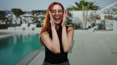 Lüks bir otel havuzunun yanında neşeli bir şekilde gülümseyen bir kadın açık hava tatil köyü ortamında kızıl saçlar, gözlükler ve modern ve ferahlatıcı bir havası olan günlük kıyafetler sergiliyor..