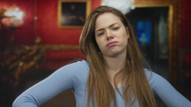 Kollarını kavuşturmuş ve dudaklarını bükmüş bir kadın sanat galerisinde çerçeveli resimler ve avizeyle duruyor..