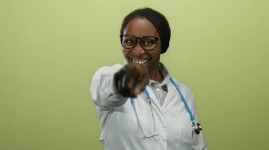 Gözlüklü ve steteskoplu kadın doktor sarı arka planda gülümsüyor, tıbbi bir ortamda kamerayı işaret ederken neşeli bir ifade sergiliyor..