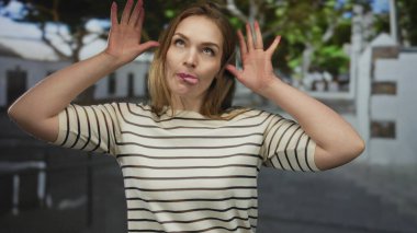 Genç beyaz kadın bir şehir caddesinde oyuncu jestlerle dil uzatır ve ellerini kaldırır..