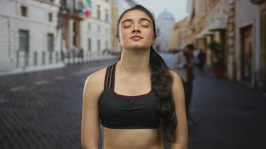 Siyah spor sutyenli genç İspanyol kadın çıplak göbeğiyle gülümsüyor kaldırım taşı sokağında gözleri kapalı, dinginlik içinde..