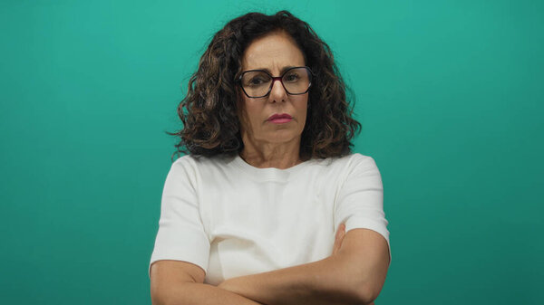 Латиноамериканка средних лет скрещивает голые руки, надевая очки в зеленой студии с серьезным хмурым взглядом; отрицание.