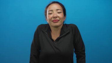 Gözleri kapalı ve ince gülümsemeli bir kadın mavi stüdyoda yüzünü ve boynunu gösteren siyah fermuarlı bluz giyen, rahat duruşu olan sakin farkındalığı olan bir kadın..