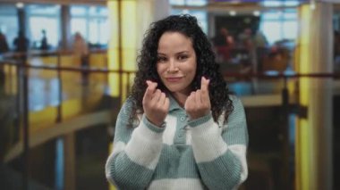 Otel lobisindeki bir kadın elleriyle para hareketi yapıyor, rahat bir kazakla güvenle gülümsüyor..