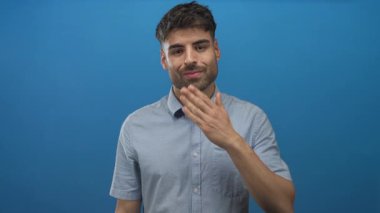 Genç İspanyol bir adam, parlak mavi bir stüdyo ortamında kameraya bir öpücük jesti için el ele tutuşur; sevgi daveti.