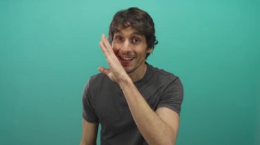 Ağızdan ağıza sarmaş dolaş genç adam gri t-shirt ile fısıldıyor stüdyoda, deniz mavisi arka planına karşı; gizlilik.