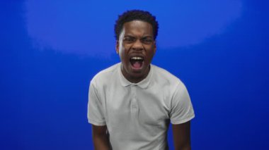 Mavi stüdyoda beyaz polo tişört giymiş, ağzı açık bağıran bir adam. Öfke gerilimi boşaltıyor..