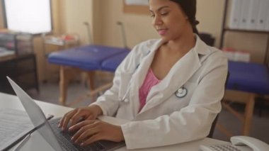 Genç Latin doktor kadın, klinik binasındaki beyaz masada steteskop ve sağlık çizelgesi ile dizüstü bilgisayarda yazı yazıyor. Verimlilik hassasiyeti sakin..