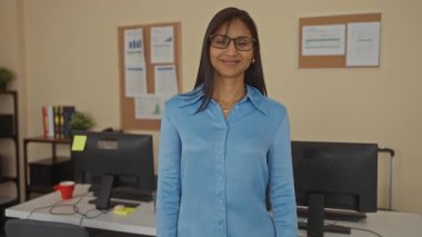 Ofis ortamında bilgisayar masasıyla gülümseyen bir kadın, duvarda tablolar, kitaplar ve bitkiler, mavi gömlek ve gözlük takan, kollar çapraz, güven ve profesyonelliği simgeleyen.