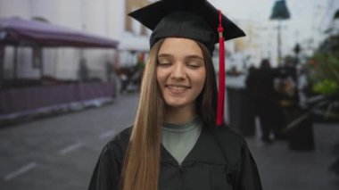 Mezun olmuş genç bir kız el oluşturma ile gülümsüyor. Meşgul bir sokakta kırmızı havan topu püskülüne imza atıyorum..