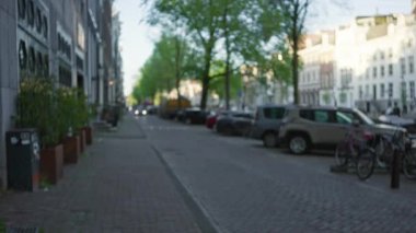 Gün ışığında park halindeki arabaların, bisikletlerin ve tarihi binaların bulunduğu bir Avrupa kentsel caddesinin odaklanmamış görüntüsü, kaldırım taşlarını kaplayan ağaçlarla kentsel bir atmosfer yakalıyor..