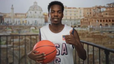 Genç Afrikalı Amerikalı adam parlak turuncu basketbol topunu tutuyor ve bulutlu gökyüzünün altındaki harabelerin yakınlarında baş parmak kaldırıyor..