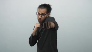 Yumrukları havada, açık gri stüdyoda gözlüklü ve sakallı genç bir İspanyol, kamera için boks pozu..