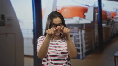 Kadın her iki işaret parmağı da güvertedeki filika ve tırabzanın yanındaki kamerayı işaret ediyor, sıradan çizgili tişörtle gülümsüyor; neşeli yaz eğlencesi.