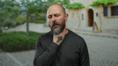 Sakallı kel bir adam şehir ortamında boğaz ağrısı çekiyor. Gün ışığında ağaçlar ve taş binalarla dolu bir şehir zemininde rahatsızlık gösteriyor..