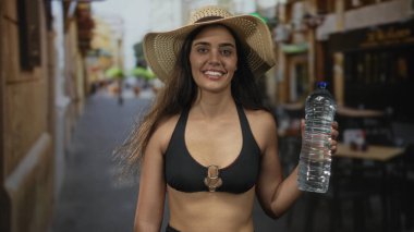Hasır şapka takan kadın dar bir sokakta parlak güneş ışığı altında geniş ve berrak bir plastik su şişesi tutuyor..