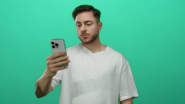 Stüdyo ortamında izole edilmiş yeşil arka planda akıllı telefon tutan sakallı genç adam gündelik kıyafetlerle modern teknoloji ve iletişim temasını gösteriyor..