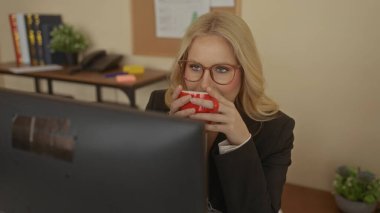 Ofisteki bir bilgisayarda çalışan, etrafı bitkilerle, kitaplarla ve ofis malzemeleriyle çevrili, kahve yudumlayan, profesyonel bir ev ortamını taşıyan gözlüklü genç bir kadın..