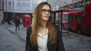 Gözlüklü ve ceketli bir kadın, kırmızı bir otobüs ve telefon kulübesinin yanındaki bir sokak boyunca bakıyor; tefekkür.