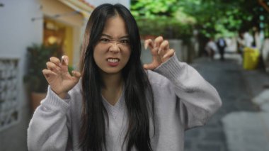 Yeşilliklerle dolu bir sokakta neşeli bir jest yapan, günlük kıyafetler giyen ve enerjik bir şekilde gülümseyen, hayat dolu bir şehir ortamında güven ve neşe saçan bir kadın..