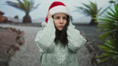 Çölde Noel Baba şapkası takan bir kadın, hayal kırıklığı içinde, palmiye ağaçlarıyla çevrili, tatil sezonunda stresi ifade ediyor..
