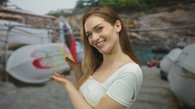 Beyaz bluzlu gülümseyen kadın güneşli sahil rıhtımında açık avuçlu beyaz kano sunar; tatil neşesi.