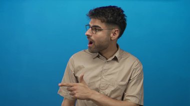 Ağzı açık ve kafası yana dönmüş bir adam mavi stüdyo duvarına karşı gözlüklü ve bej gömlekli; sürpriz.