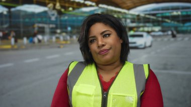 Yansıtıcı yelek giyen genç İspanyol kadın havaalanı terminalinin önünde duruyor, dalgın bir görünüm sergiliyor, bulanık bir geçmişi var ve yoğun havaalanı ortamını vurguluyor..